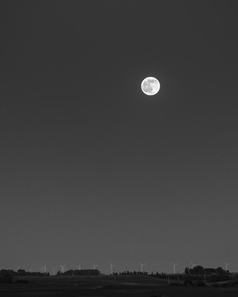 Black and white photograph of the moon over distant wind turbines at dusk